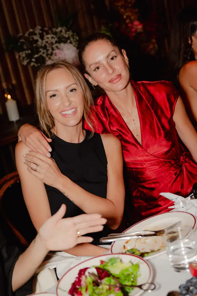 Two women are seated closely at a dinner table, smiling warmly at the camera. The woman on the left, wearing a black top, has her arm around the woman on the right, who is dressed in a vibrant red patterned dress and a delicate gold necklace. Plates of food, including salads, and elegant floral arrangements are visible on the table and in the softly lit background, indicating a social event or dinner.