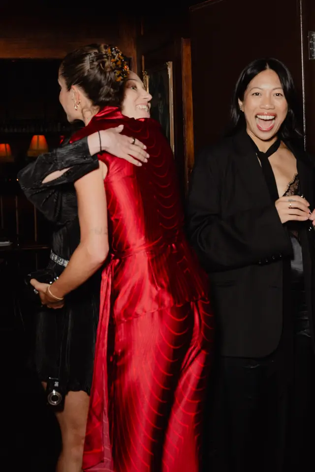 Three women are captured in a candid moment at a dimly lit indoor event. Two women are embracing, with one wearing a vibrant red patterned dress and the other a black pleated dress. A third woman, wearing a black blazer and tie over a lace top, stands to the right, laughing with her mouth open.
