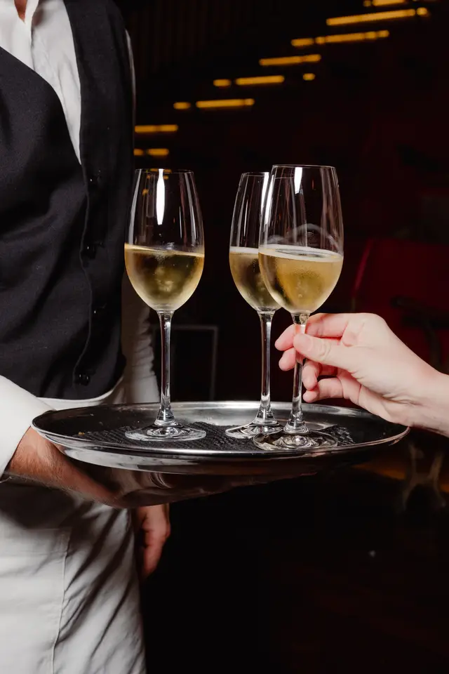 A close-up shot captures a person's hand reaching for a glass of sparkling wine from a silver tray. The tray is held by a waiter, whose torso is visible in a white shirt and black vest. Three glasses filled with a golden, bubbly liquid are presented on the tray against a dark, softly lit background.