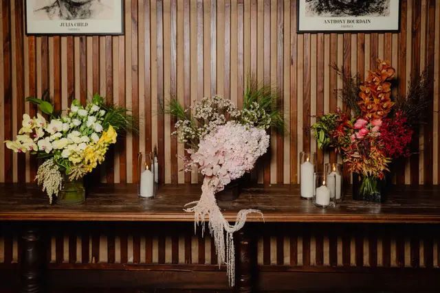 Three elaborate floral arrangements are displayed on a dark wooden table against a slatted wooden wall. Each arrangement features a distinct color palette, flanked by lit pillar candles in clear glass holders. Two framed black and white prints, one of Julia Child and one of Anthony Bourdain, hang above the table.