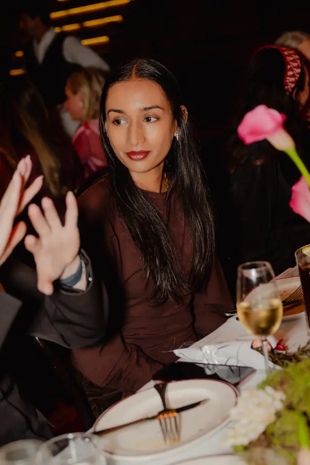 A candid, vertical shot of a woman with long dark hair and a brown top, seated at a table during an evening event. She is looking to her right, with a wine glass, a plate with cutlery, and pink calla lilies visible on the table in the foreground. The background features blurred figures of other attendees and warm, out-of-focus lights.
