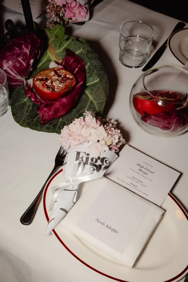 A close-up, overhead shot of an elegant table setting for the 'Fig & Bloom High Floral Design' event. The setting features a white plate with a red rim, a menu card, a place card, cutlery, and a small bouquet of light pink flowers wrapped in branded paper. Decorative elements like radicchio, pomegranate halves, and grapes are artfully arranged on the white tablecloth.