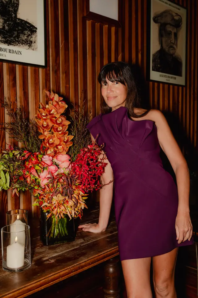 A woman with dark hair and a purple one-shoulder dress smiles while leaning on a rustic wooden table. To her left, a large, vibrant floral arrangement in a dark vase features orange orchids, pink ranunculus, and various red and green foliage. Two lit white candles in glass holders are also visible on the table, with a wooden paneled wall and framed portraits in the background.