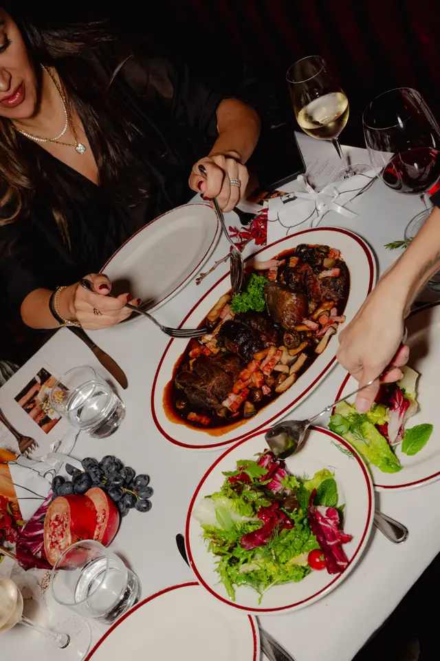 An overhead shot captures a dining scene with multiple hands reaching for food and drinks on a white tablecloth. A large platter of braised meat with mushrooms and bacon is central, flanked by plates of fresh green salad. Glasses of red wine, white wine, and water are also visible, alongside decorative elements like grapes and a sliced pomegranate.