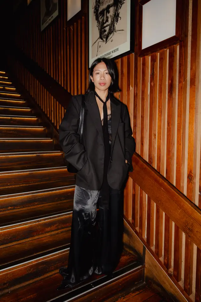 A woman in a black blazer and flowing black pants stands on a dimly lit wooden staircase, looking directly at the camera. The staircase features illuminated steps and a wooden handrail, while the background consists of a wooden slat wall adorned with framed artwork. The overall scene suggests an elegant indoor event or venue.