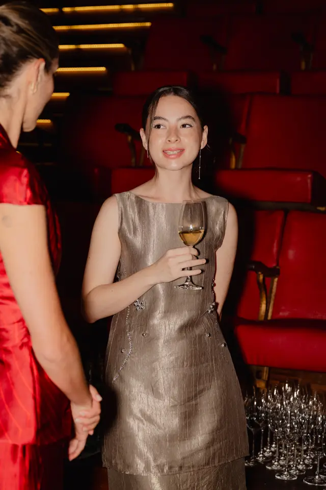 A young woman in a shimmering champagne-colored dress, adorned with sparkling embellishments, smiles while holding a glass of white wine. She is looking towards another person, whose back is partially visible in a red garment. The background features rows of plush red theater-style seats and a collection of empty wine glasses, suggesting an elegant social event.