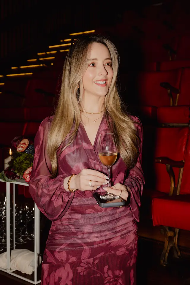 A smiling woman with long blonde hair, wearing a maroon patterned dress, holds a wine glass and a smartphone. She is positioned in front of rows of red theater seats, with a floral and fruit arrangement visible to her left. The scene suggests an elegant evening event.