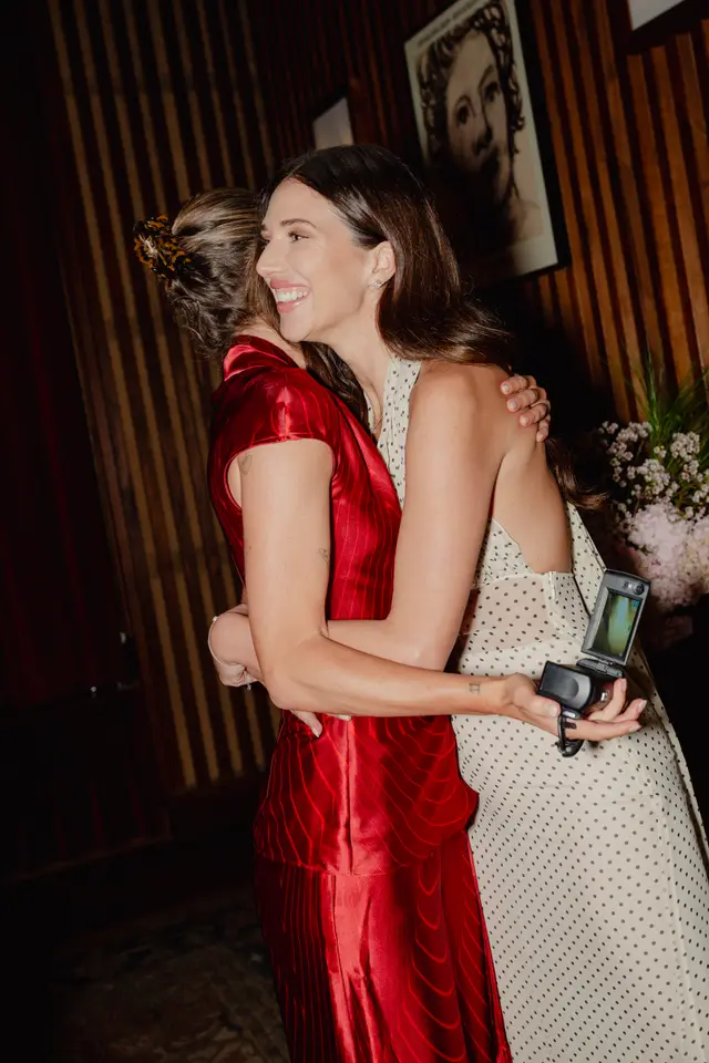 Two women are captured in a warm embrace, one smiling broadly while holding a camcorder. They are positioned in an indoor setting with dark wooden paneling and a framed artwork in the background, alongside a floral arrangement.