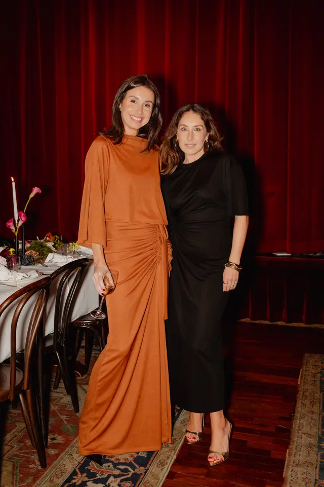 Two women stand smiling in an elegant indoor setting, posing for a photo. The woman on the left wears a rust-colored long dress and holds a wine glass, while the woman on the right wears a black dress. Behind them, a set dining table with floral arrangements, candles, and branded napkins is visible against a rich red curtain backdrop.