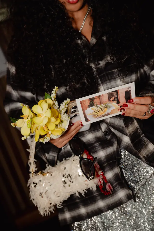 A close-up shot captures a person holding a small bouquet of yellow orchids and white bell flowers, a smartphone, and a printed photo. The person is adorned with red nail polish, rings, and a necklace, wearing a plaid shirt and a sequined skirt. The image focuses on the details of the hands and accessories, suggesting a festive or celebratory event.