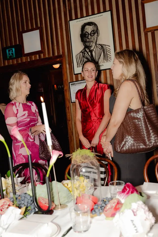 Three women are gathered around a beautifully decorated table at an indoor event. The foreground features an elaborate floral and fruit centerpiece with lit candles, while the background shows dark wood-paneled walls and framed artwork. The atmosphere appears social and elegant, with warm lighting.