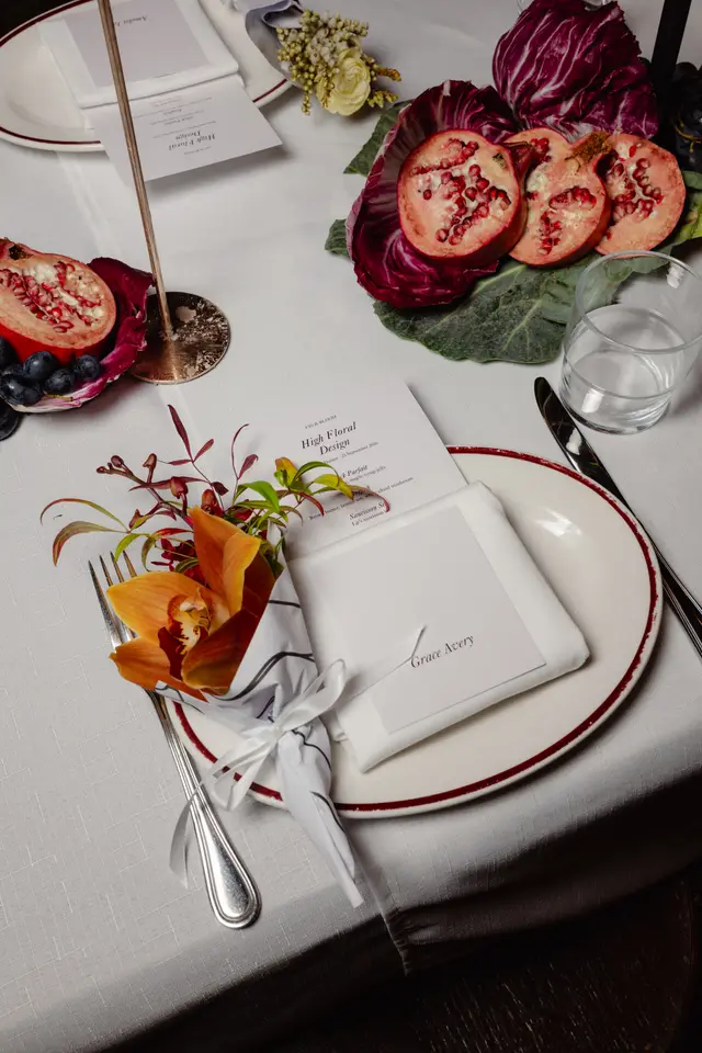 A detailed overhead shot of an elegantly set table for a 'High Floral Design' event. The setting features individual place settings with personalized name cards, intricate floral arrangements, and decorative elements like halved pomegranates, radicchio, and fresh berries. The composition highlights a blend of rich colors and textures against a crisp white tablecloth, suggesting a sophisticated and autumnal theme.