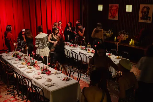 An overhead shot captures a lively indoor event with multiple guests mingling around a long dining table. The table is elegantly set with a white tablecloth, place settings, candles, and elaborate floral and fruit arrangements. Red curtains line one wall, contributing to the warm, intimate ambiance created by the dim lighting and candlelight.