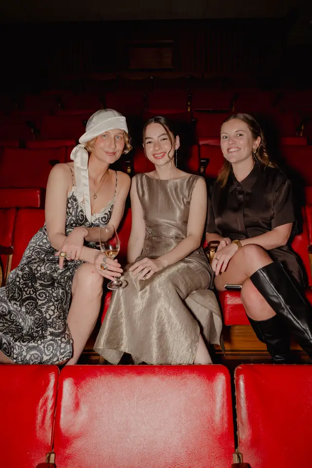 Three women are seated in a row of red upholstered theater seats, smiling and looking towards the camera. They are dressed in elegant attire, with two holding wine glasses. The background is dark, highlighting the subjects and the vibrant red of the seating.