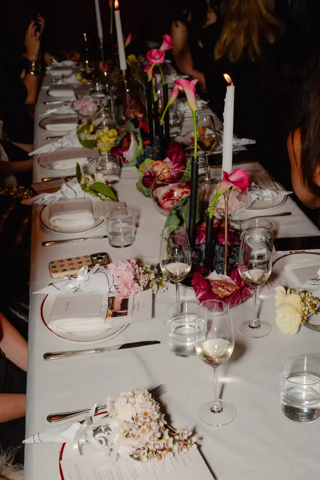 An elegant dinner table is set for an event, featuring elaborate floral and fruit arrangements as centerpieces. White tablecloths, formal place settings, and lit candles create a warm and sophisticated atmosphere. The image captures a section of a long table, highlighting the detailed decor and the presence of guests.
