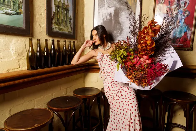 A woman in a white and red polka dot dress, wearing a pearl necklace, leans against a wooden shelf while holding a large, vibrant bouquet of flowers. The setting is a warm-toned interior, possibly a bar or restaurant, featuring a brick wall, framed artwork, and wooden bar stools. The woman gazes to her right, creating an elegant and contemplative mood.