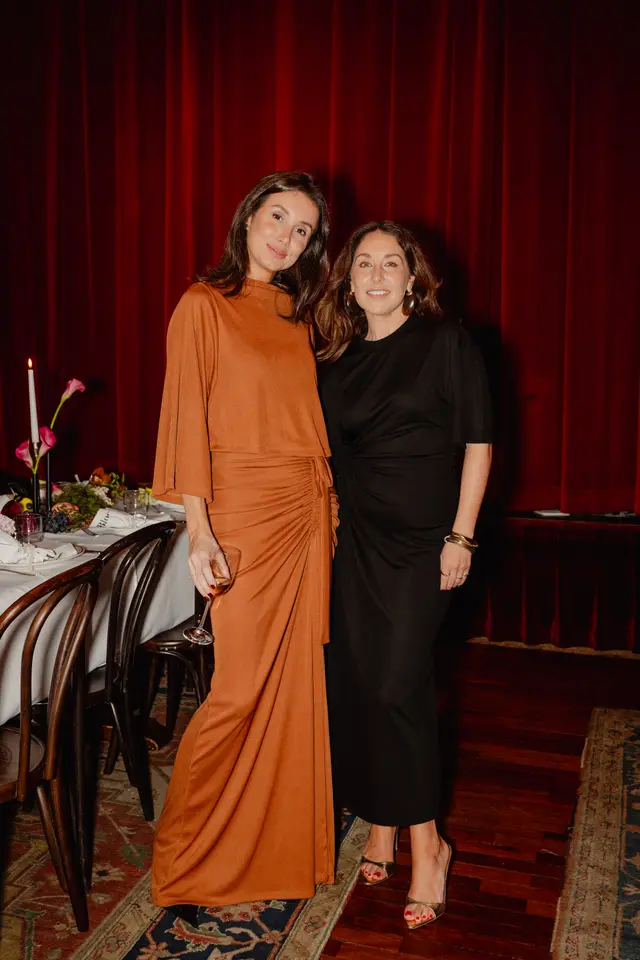 Two women, dressed in elegant brown and black dresses, stand smiling in front of rich red curtains. A beautifully set dinner table with a white tablecloth, floral arrangements, a lit candle, and grapes is visible to their left. The scene captures a sophisticated evening event.