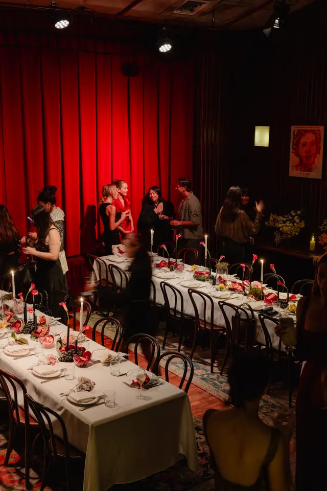 The image captures an elegant indoor event with multiple guests mingling around long, white-clothed dining tables. The tables are elaborately decorated with floral arrangements, candles, and fruit, set against a backdrop of rich red curtains and dark wooden paneling. Stage lighting illuminates the festive atmosphere, with some guests in motion, contributing to a lively yet sophisticated ambiance.