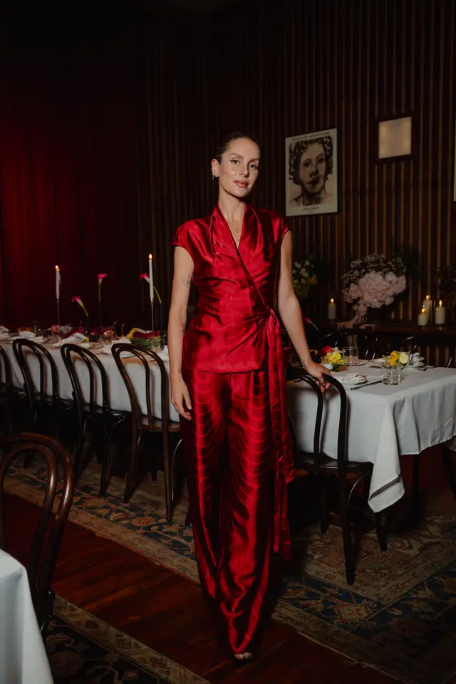 A full-length shot of a woman in a striking red satin wrap top and matching wide-leg pants, standing beside a long, elegantly set dinner table. The room features dark wood paneling, a patterned rug, and soft, warm lighting from candles and ambient sources. Various floral arrangements adorn the table and a side console, creating an intimate and sophisticated atmosphere.