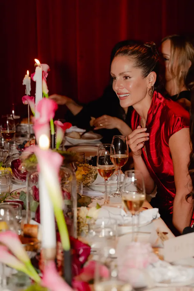 A woman in a red patterned top smiles while seated at a beautifully decorated dinner table. The table is adorned with lit candles, pink calla lilies, and various food items, creating a warm and elegant atmosphere. Other guests are visible, blurred in the background.