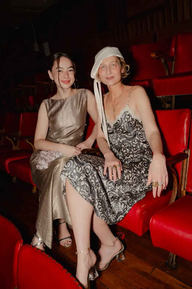 Two women are seated side-by-side in a row of red theater seats, looking towards the viewer with slight smiles. One woman wears a shimmering gold sleeveless dress, while the other wears a black and white patterned slip dress with white lace trim and a white cloche-style hat. The setting features dark wooden floors and rows of empty red seats in the background.