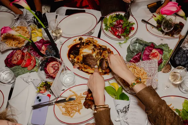An overhead shot captures a bustling dinner table laden with various dishes, drinks, and floral arrangements. Multiple hands are visible, some reaching for food and interacting with the table setting, suggesting a lively and social dining experience. The table is set with white tablecloths, formal plates, and an array of colorful food items.