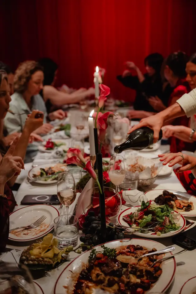 A warm, intimate shot captures multiple people dining at a long table, richly decorated with various dishes, wine glasses, lit candles, and floral arrangements. A hand is visible pouring wine into a glass, while other guests are engaged in eating and conversation. The background features deep red curtains, contributing to a luxurious and festive atmosphere.
