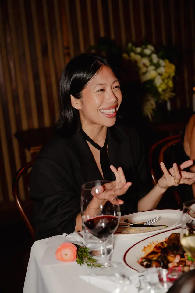 A smiling woman with dark hair, wearing a black blazer and tie, is seated at a white-clothed dinner table, gesturing with her hands. The table is set with a glass of red wine, plates of food, and a small pink ranunculus with a sprig of rosemary. A blurred floral arrangement and a dark, striped wall are visible in the background, suggesting an elegant event.