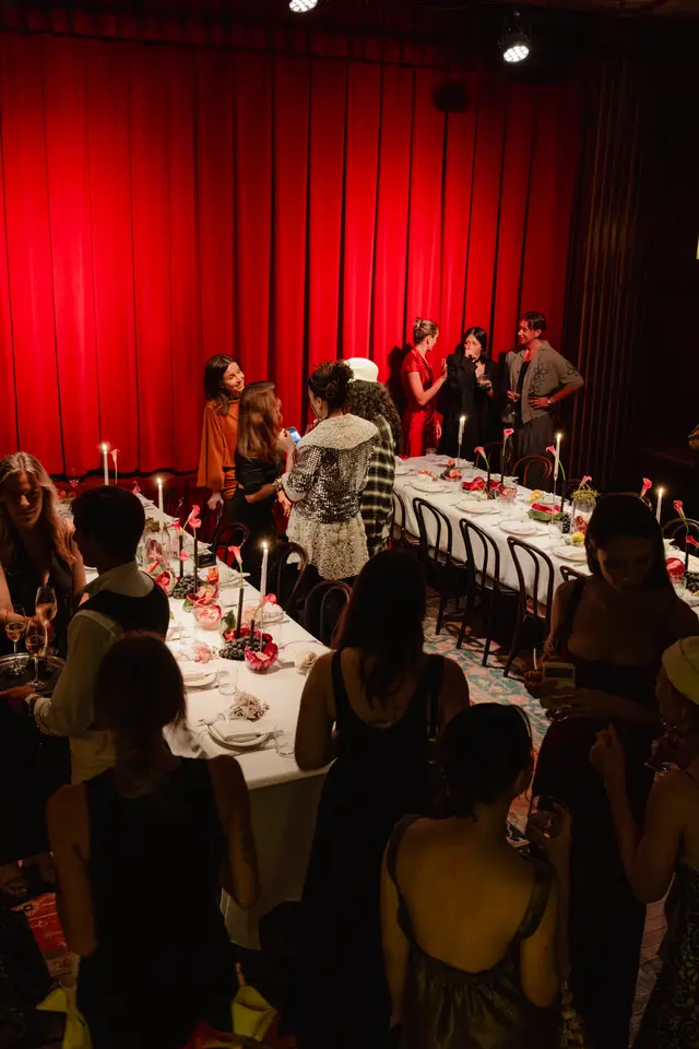 The image captures an indoor evening event with multiple guests gathered around long dining tables. The setting features a vibrant red curtain backdrop, tables adorned with white tablecloths, lit candles, and floral arrangements. People are seen socializing, holding drinks, and interacting in a warm, intimate atmosphere.