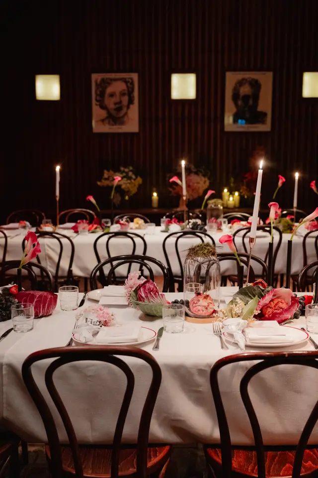 A beautifully set dinner table for an event, featuring white tablecloths, dark wooden chairs, and elaborate floral and fruit centerpieces. The dimly lit room creates an intimate atmosphere, with glowing candles providing warm light. Framed artwork and subtle wall sconces are visible on the dark, textured wall in the background.