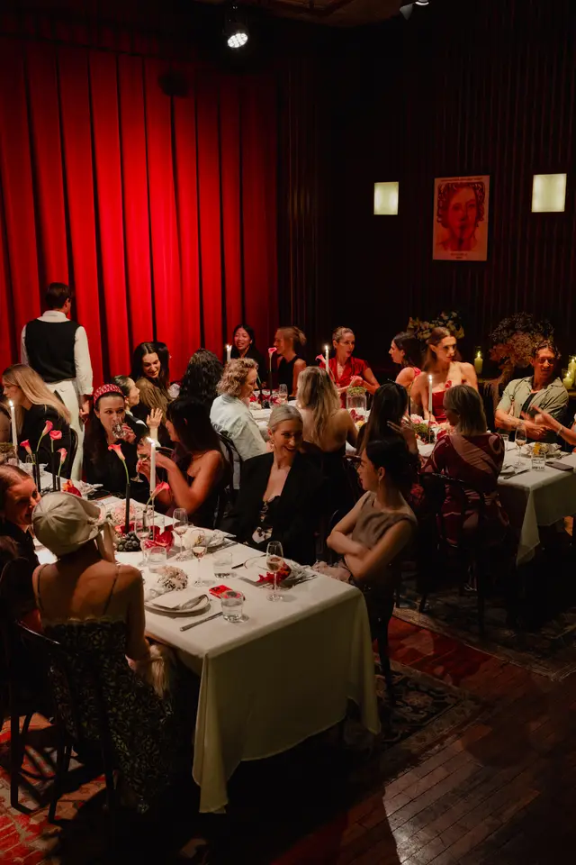 A vibrant, low-light photograph captures a lively dinner event with multiple guests seated at tables adorned with white tablecloths, candles, and floral arrangements. A waiter stands near a prominent red curtain backdrop, while guests engage in conversation and enjoy the evening. The setting suggests an intimate and festive gathering.