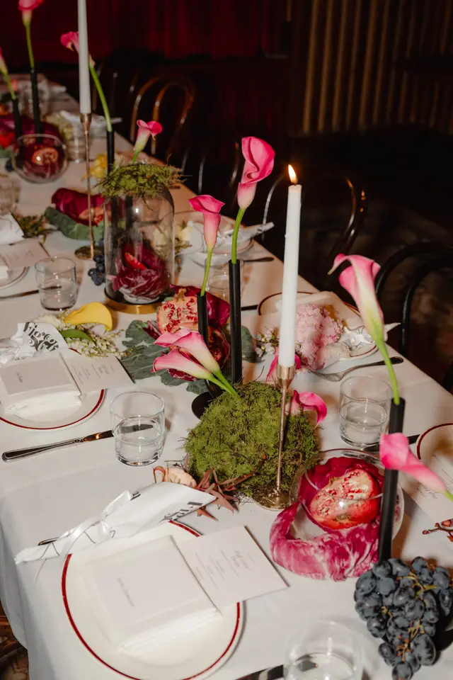 A beautifully arranged long dining table is set for an event, featuring multiple pink calla lilies in black and gold holders, interspersed with lit candles. Various natural elements like halved pomegranates, bunches of dark grapes, and green moss are used as decor. Individual place settings with white plates, folded napkins, and menu cards are visible, suggesting an elegant dinner or launch event in a dimly lit, warm interior.