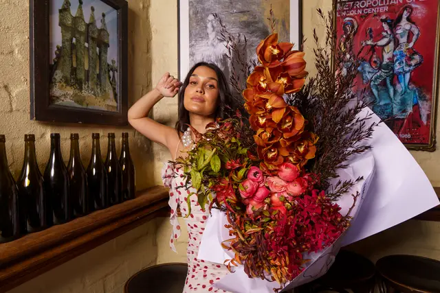 A woman in a white dress with red polka dots holds a large, vibrant bouquet of orange, pink, and red flowers wrapped in white paper. She poses against a textured light brown wall, looking directly at the camera with a subtle smile. The background features framed artwork, a wooden shelf with dark bottles, and dark wooden stools, creating a warm and art-filled ambiance.