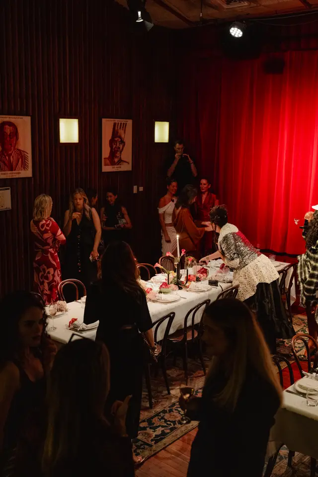 An indoor event scene captured from an elevated perspective, showing multiple guests mingling around a long white-clothed dining table. The room features dark wood paneling, red curtains, and warm, dim lighting, creating an intimate and festive atmosphere. Floral arrangements, candles, and place settings adorn the table, suggesting a special occasion or dinner event.