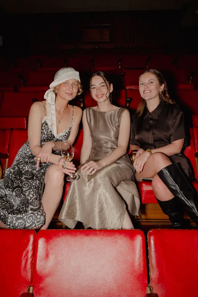Three women are seated in a row of red theater seats, smiling towards the camera. They are dressed in formal or semi-formal attire, with one holding a wine glass. The background is dark, showing more rows of empty red seats in an auditorium setting.