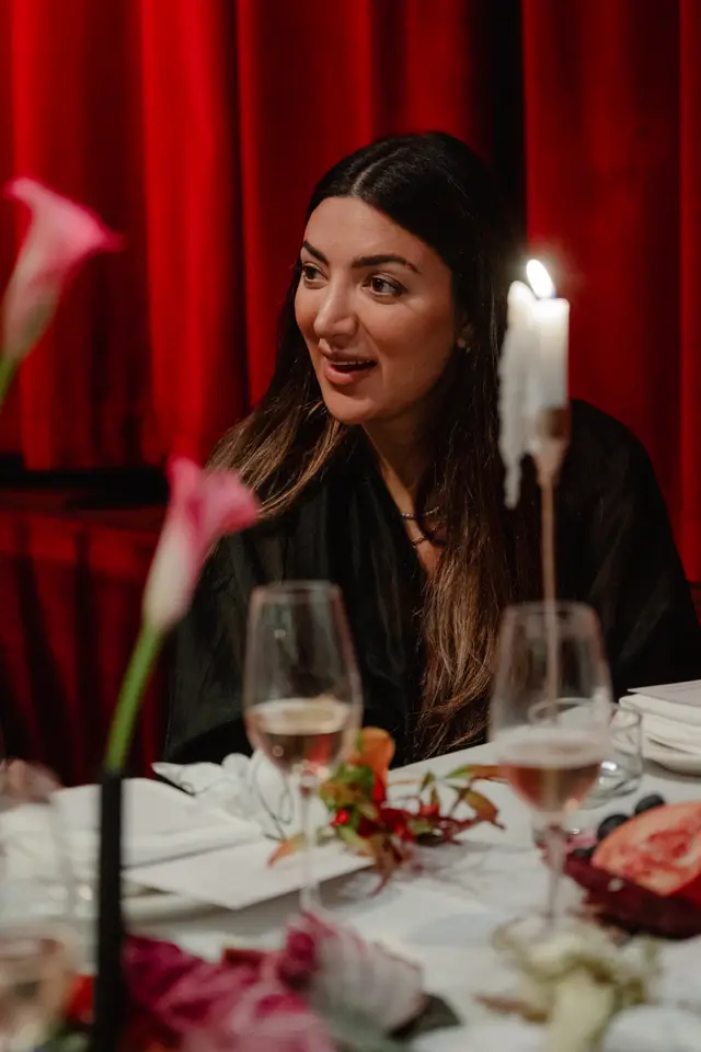 A close-up shot of a woman with long dark hair, smiling and looking to the side, seated at a table during an event. The background features rich red curtains, and the table is adorned with a lit candle, wine glasses, and floral arrangements. The lighting is warm and intimate, highlighting the woman and the table setting.