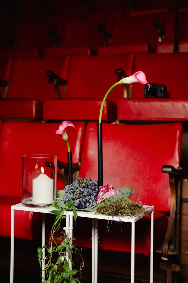 A styled shot featuring a white console table adorned with two pink calla lilies in black vases, a lit white candle in a glass hurricane, a bunch of dark grapes, radicchio, and moss. Green foliage trails from the table's base. The arrangement is set against a blurred background of empty red theater seats, creating a dramatic and elegant atmosphere.