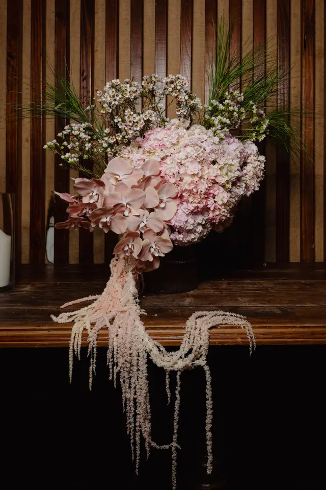 A large, elaborate floral arrangement featuring light pink Phalaenopsis orchids, clusters of pink hydrangeas, and delicate white wax flowers, accented with green spiky foliage. Long, trailing light pink amaranthus cascades down the front of the arrangement, which is set in a dark, ribbed vase on a rustic wooden surface. The background consists of a wall with vertical dark wood strips against a lighter, textured fabric.