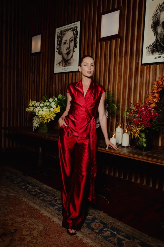 A woman in a striking red satin wrap top and matching wide-leg pants stands elegantly beside a dark wooden table. The table is adorned with lush floral arrangements in yellow, white, red, and orange tones, alongside several lit candles in glass holders. The background features dark wooden vertical paneling with framed artwork and wall sconces, while a patterned rug covers the floor.