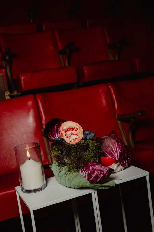 A still life composition featuring a lit white candle in a glass holder and a dramatic arrangement of a halved pomegranate, dark grapes, radicchio, and moss, all resting on a white side table. The scene is set against a blurred background of plush red theater seats in a dimly lit space.