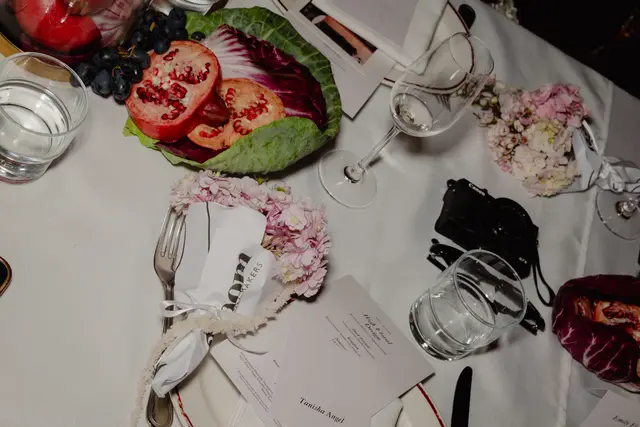 A top-down view of an elegantly set table at an event, featuring a white tablecloth adorned with various elements. Fresh pomegranates, grapes, and leafy greens are arranged alongside delicate pink floral bouquets, clear glassware, and silver cutlery. Event menus and place cards are also visible, with a black camera resting on the table.