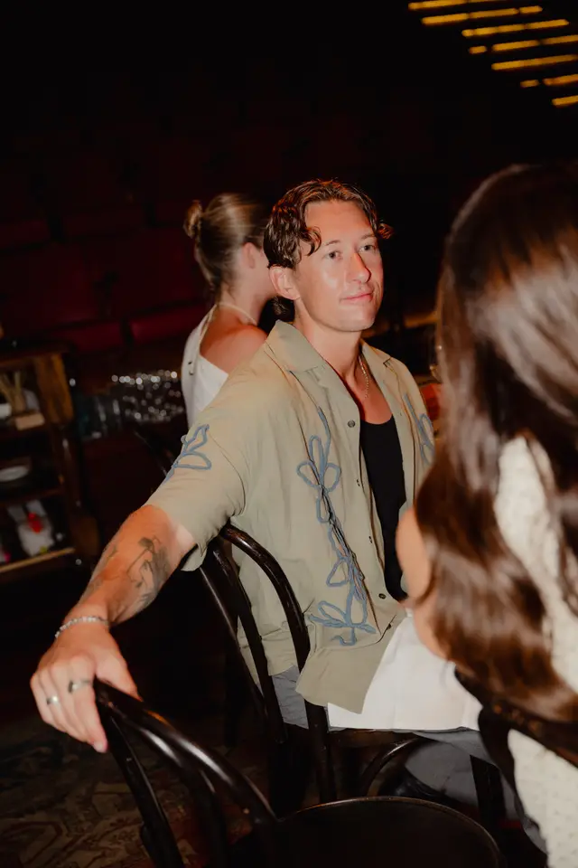 A candid, medium shot captures a man with curly hair, wearing a light olive green shirt with blue embroidered floral patterns, seated at an event. He looks to the right with a subtle smile, while other attendees are blurred in the background. The scene is dimly lit, suggesting an intimate indoor gathering with warm lighting.
