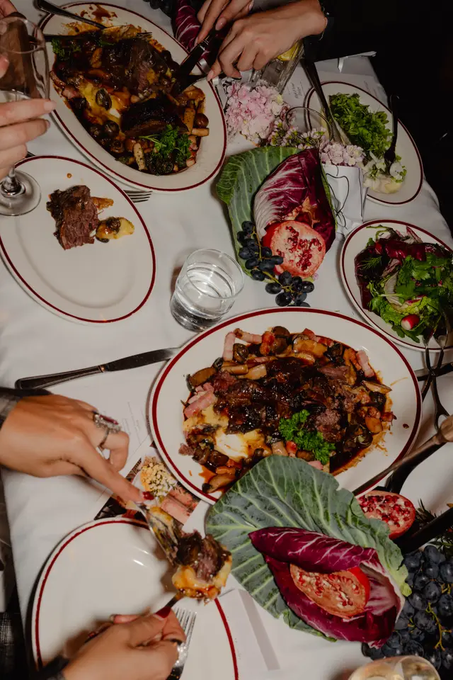 A top-down view captures a lavish dining table set for an event, featuring multiple plates of rich food, fresh salads, and decorative elements. Several hands are visible, actively engaging with the dishes and drinks, suggesting a vibrant social gathering. The table is adorned with a white tablecloth, and the scene is rich with dark meats, vibrant greens, and colorful fruits and flowers.