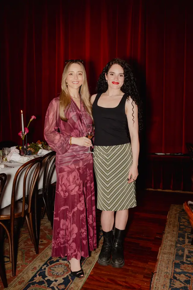 Two women stand smiling, posing for a full-length photo at an indoor event. One woman wears a long, flowing floral dress and holds a wine glass, while the other wears a black tank top and a striped midi skirt. In the background, a set dining table with floral arrangements and candles is visible, against a backdrop of dark red velvet curtains.