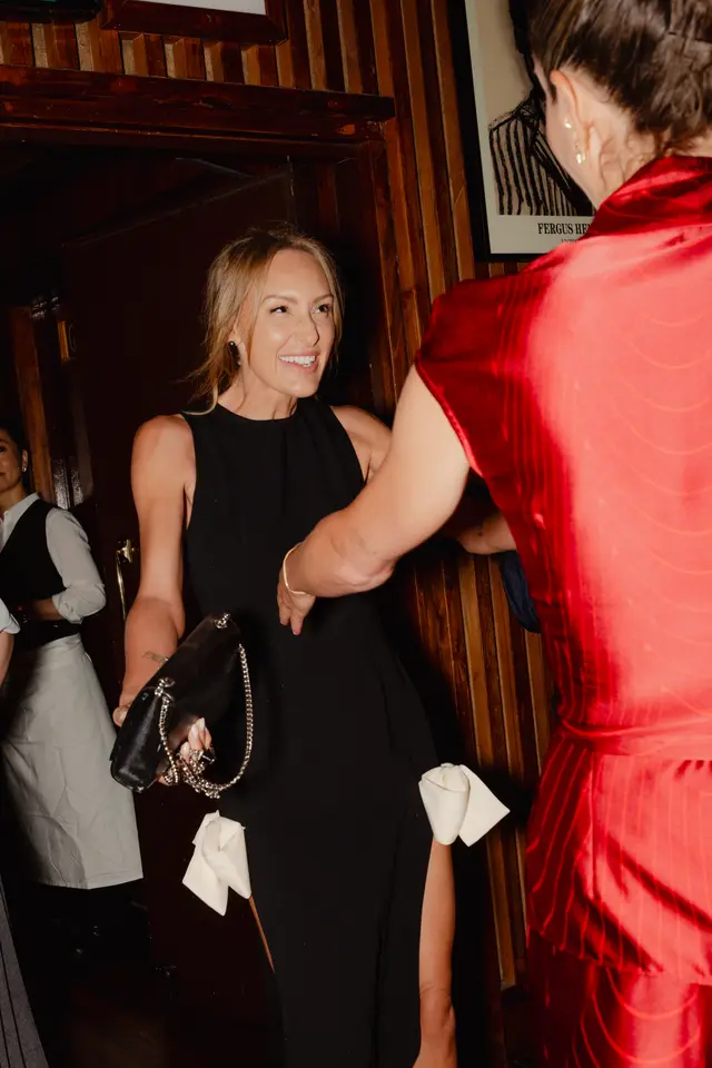A candid, vertical shot captures a smiling woman in a black sleeveless dress, holding a black clutch with a silver chain, as she interacts with another person in a red patterned outfit. The background features dark wood paneling and a doorway, suggesting an indoor event or restaurant setting. A third person, possibly a server, is partially visible on the left.