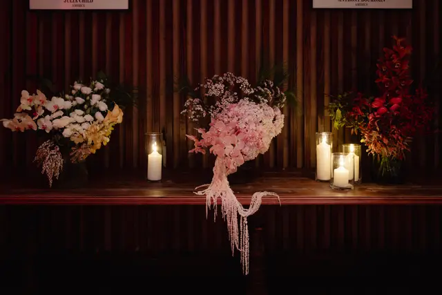 The image features three elaborate floral arrangements displayed on a dark, polished wooden table, illuminated by the warm glow of multiple candles in glass hurricane lamps. The background consists of a dark wooden slatted wall, creating an intimate and sophisticated ambiance. The arrangements showcase a variety of flowers in shades of white, cream, yellow, pink, and deep red, with lush green foliage.