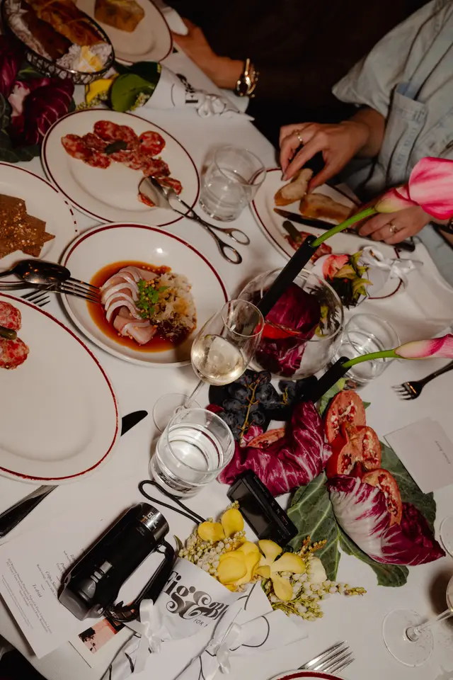 An overhead shot captures a beautifully set dinner table adorned with various dishes, drinks, and floral arrangements. Hands are visible reaching for food and holding a glass, suggesting an active dining experience. The scene is rich with details, from gourmet food plates to decorative elements and personal items.