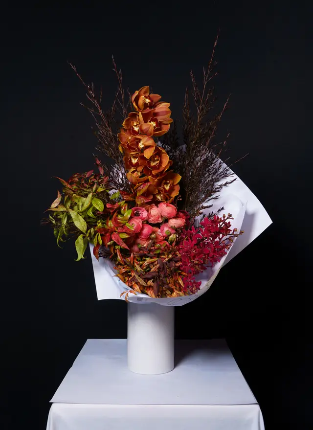 A vibrant, high-contrast studio shot of a large floral bouquet wrapped in white paper. The arrangement features a rich mix of orange orchids, coral ranunculus, and various green, red, and brown foliage. It is presented on a white cylindrical stand atop a white draped surface against a dark, plain background.