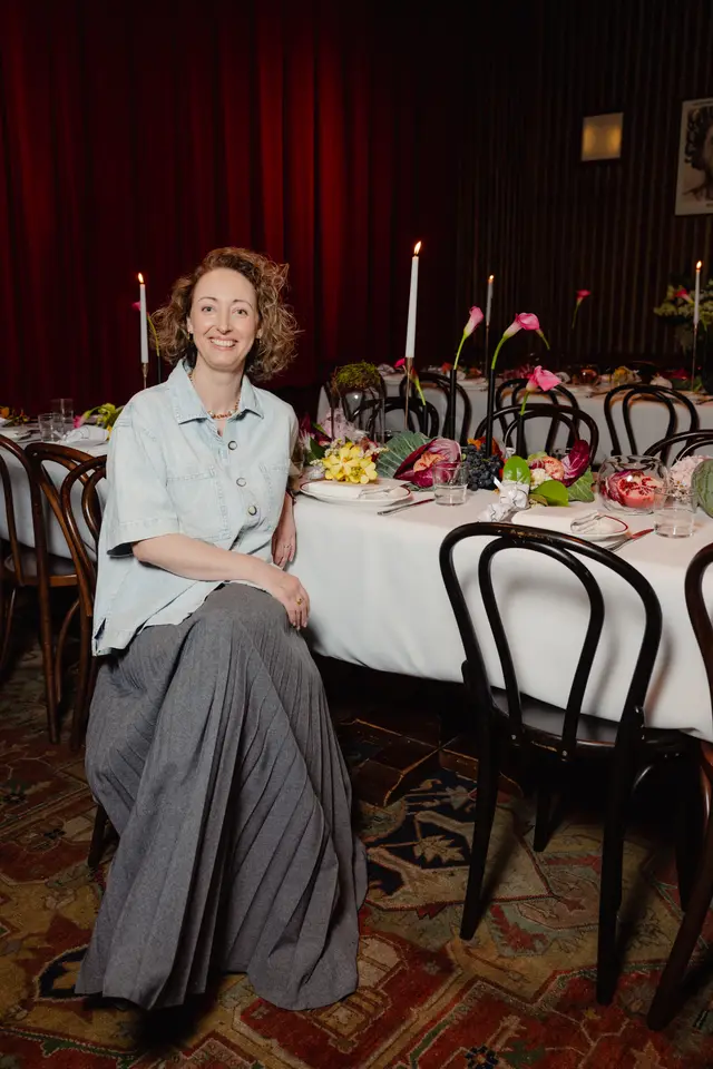 A smiling woman with curly hair is seated at a long, elegantly decorated dinner table, looking directly at the camera. The table is adorned with a white tablecloth, dark wooden chairs, and elaborate centerpieces featuring pink calla lilies, yellow orchids, grapes, pomegranates, and radicchio. The background includes dark red curtains and a richly patterned rug, suggesting a sophisticated event setting.
