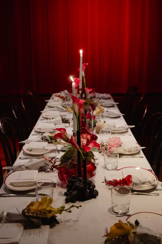 A long dining table is elegantly set for an event, viewed longitudinally from one end. The table features multiple place settings with white plates and red rims, cutlery, and clear glasses. Elaborate centerpieces include white taper candles, red calla lilies, pink carnations, green moss, and dark grapes, all set against a dramatic red curtain backdrop.